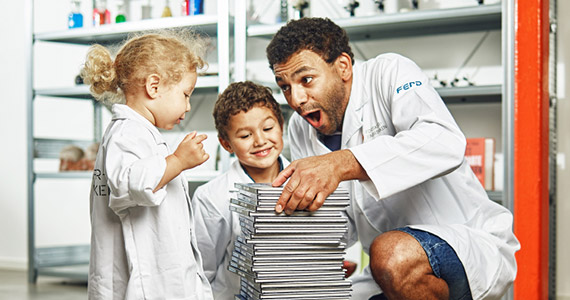 Shows a father and his two children, all wearing lab coats and excited about maths